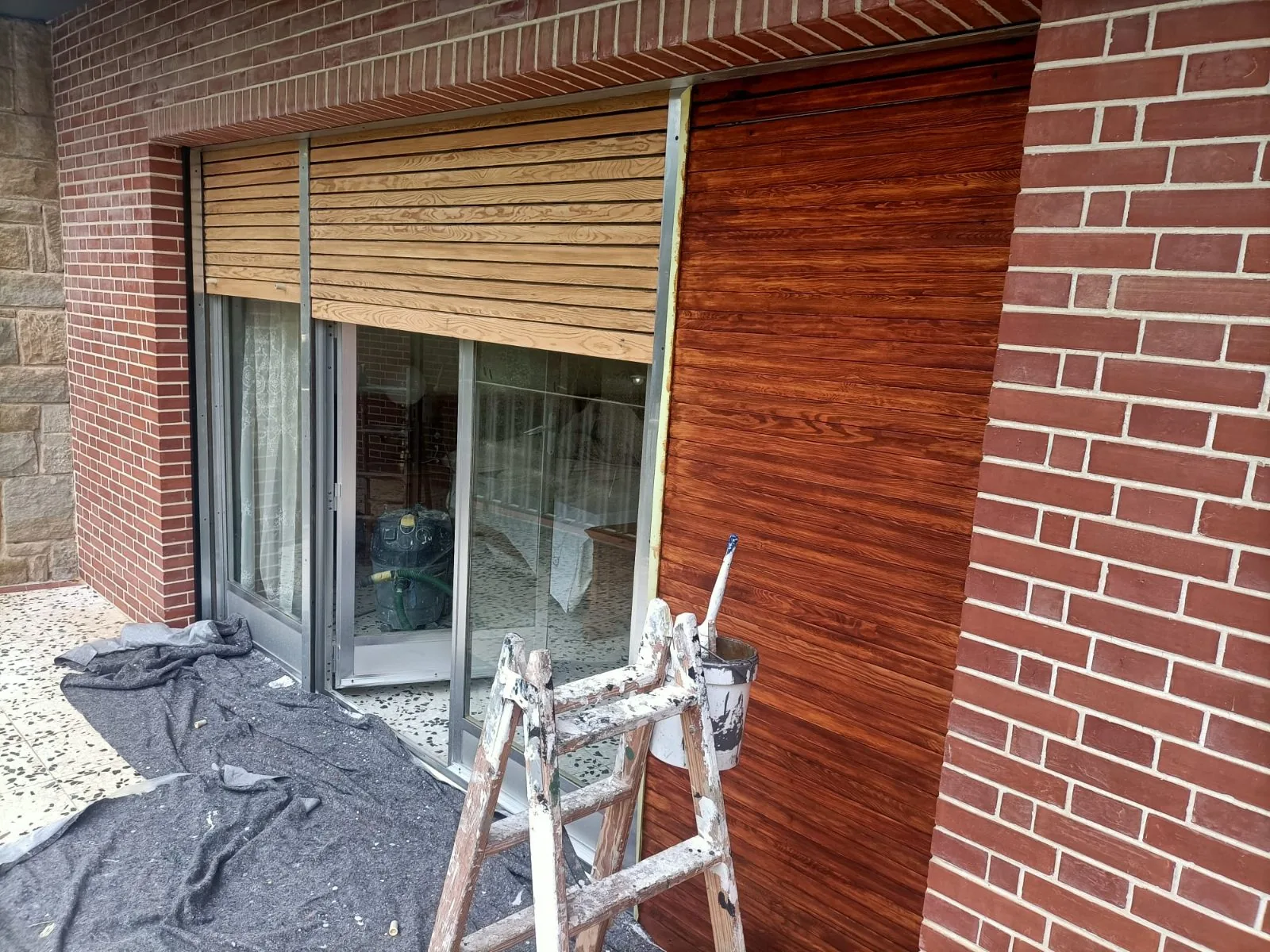 Pared exterior con acabado imitación madera junto a persiana, con escalera de pintor