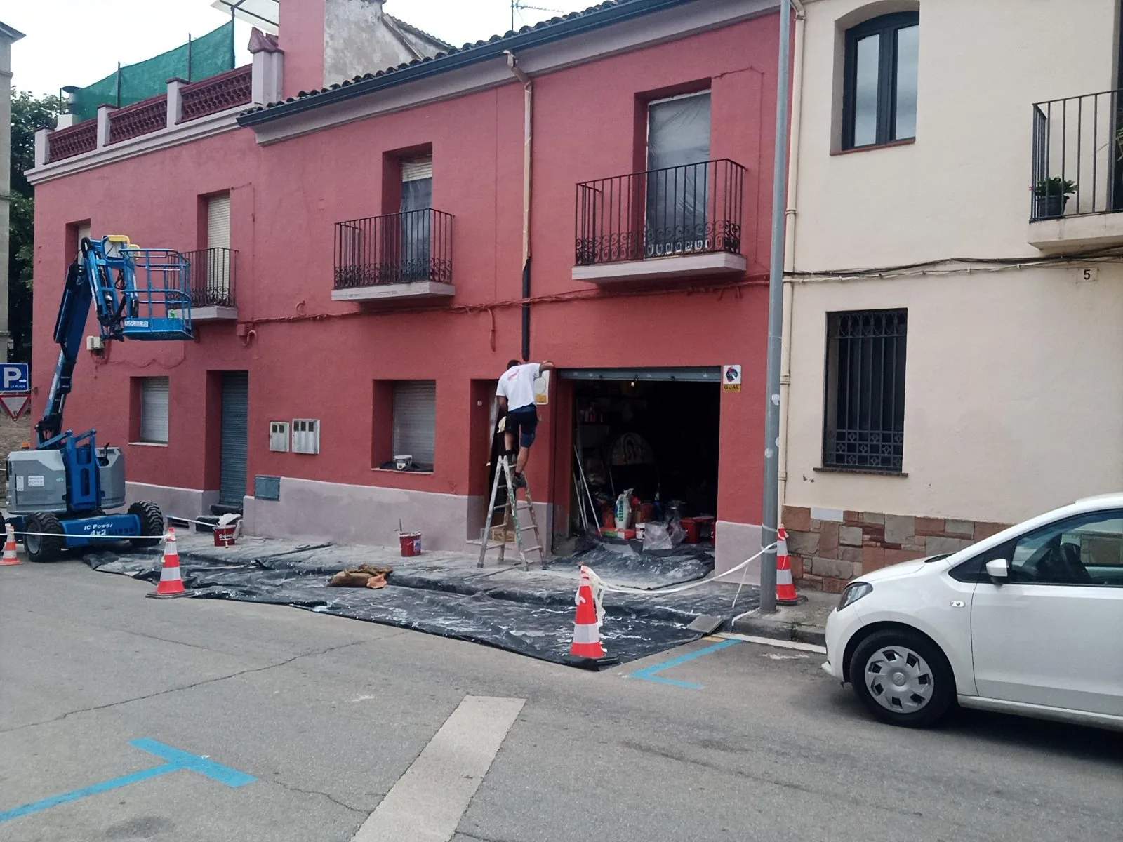 Pintor en escalera pintando una fachada roja con plataforma elevadora y la calle protegida